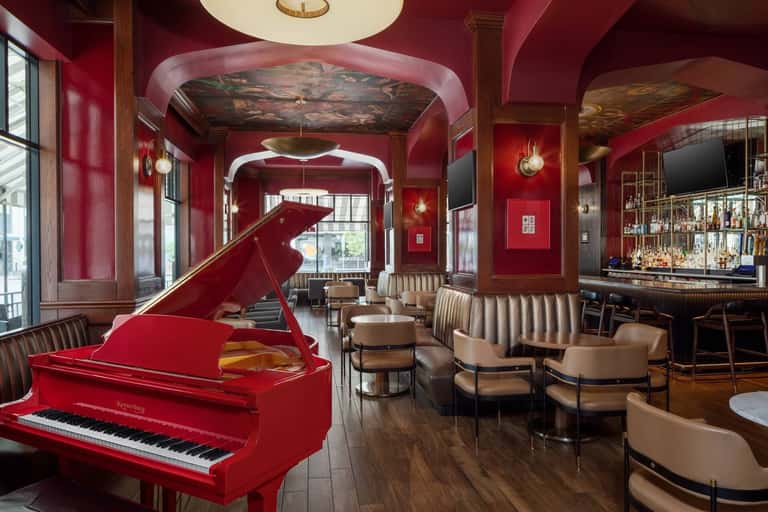 Elegant piano lounge with bright red grand piano, cream armchairs, bar area, and ornate architectural details