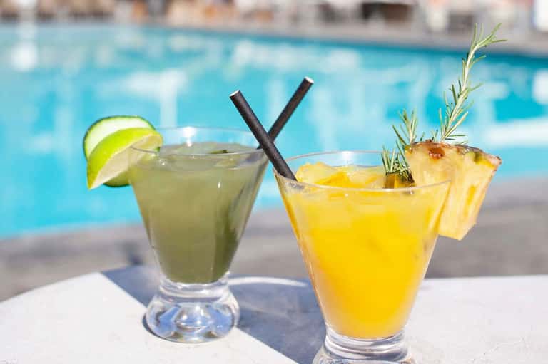 Two tropical cocktails by pool—lime mojito and pineapple drink with garnishes and straws