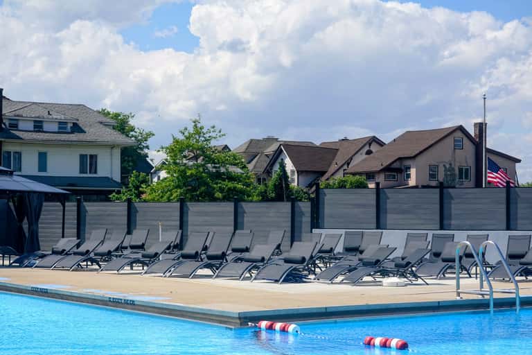 Resort pool with rows of lounge chairs, privacy fence, and residential homes in background
