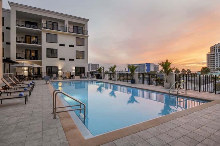 Resort pool at sunset with multi-story white building, palm trees, and city skyline