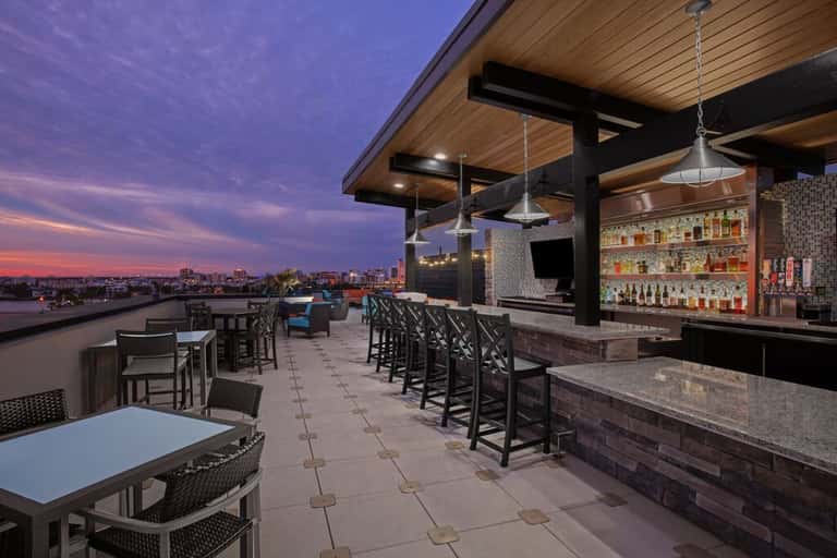 Rooftop bar with full liquor display, comfortable seating, and purple sunset cityscape views