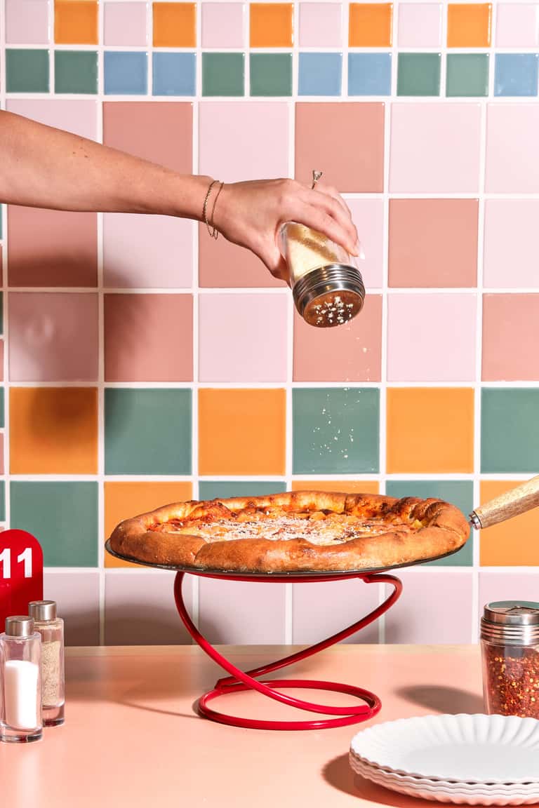 Hand sprinkling salt on fresh pizza over colorful tiled background with drink visible