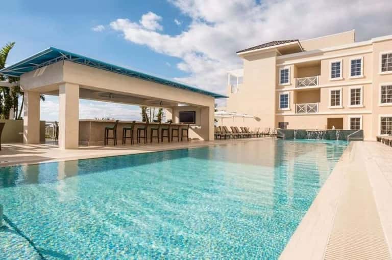 Resort pool with modern poolside bar pavilion, lounge chairs, and cream-colored building with tropical landscaping