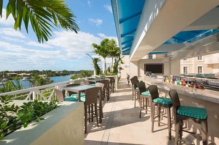 Waterfront terrace dining area with bar seating, tables, and views of the water and palm trees