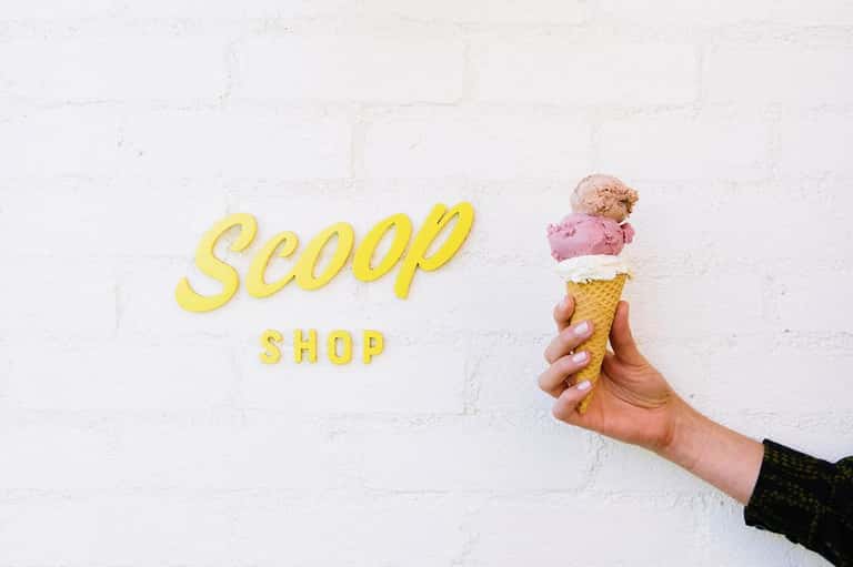Hand holding three-scoop ice cream cone in front of yellow Scoop Shop sign on white brick wall