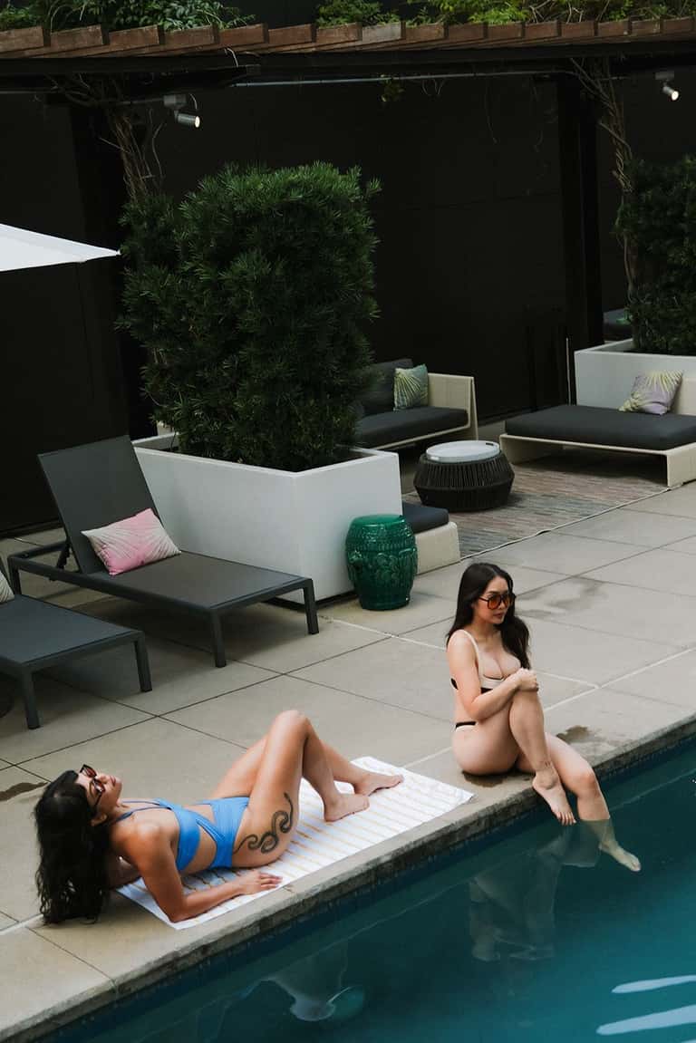 Two women relaxing on white towels by modern resort pool with lounge chairs