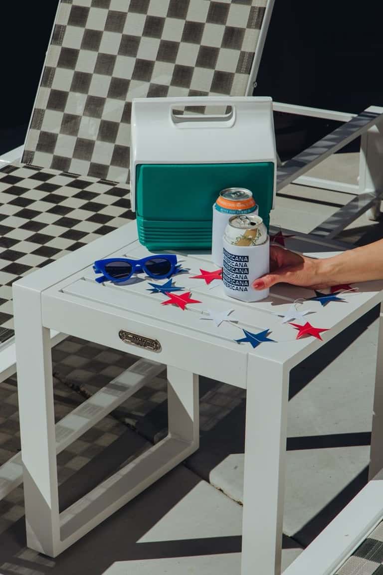 Casual outdoor seating with cooler, sunglasses, and beverage on white table with checkered pattern