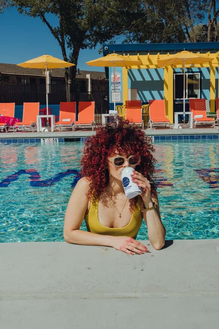 Woman in yellow swimsuit at pool edge with curly red hair, white sunglasses, holding drink with yellow umbrellas and orange loungers visible