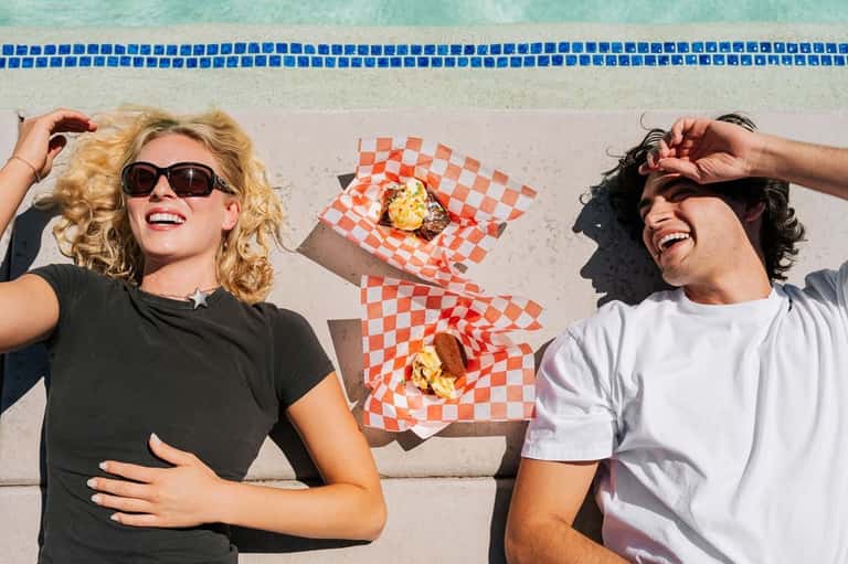 Two people in casual clothing lying on pool deck with red checkered food wrapper and items between them, pool visible behind