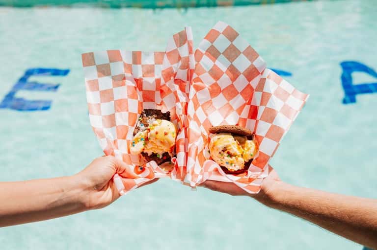 Two hands clinking burgers wrapped in red and white checkered paper over pool water with blue lane dividers