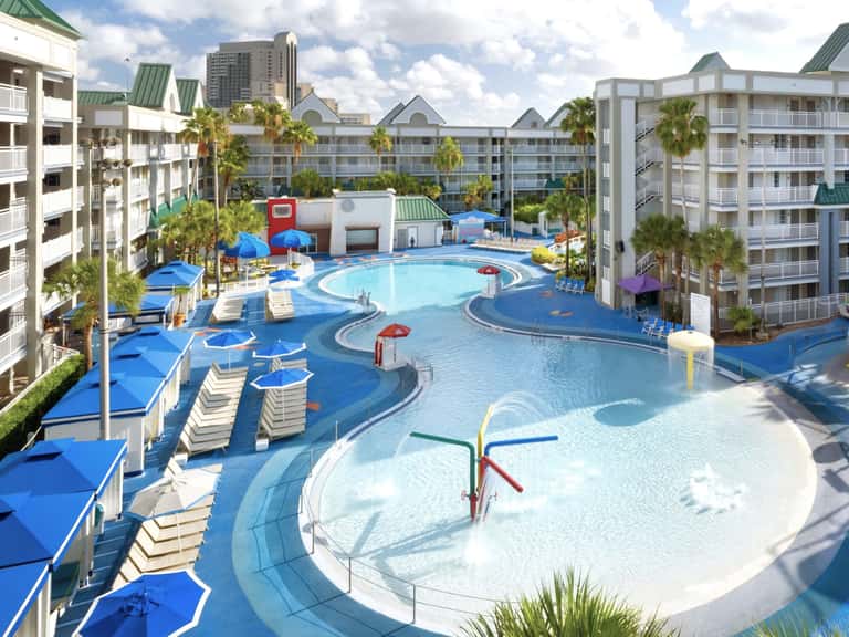 Expansive resort pool complex with blue water, loungers, umbrellas, and multi-story apartment buildings surrounding the area
