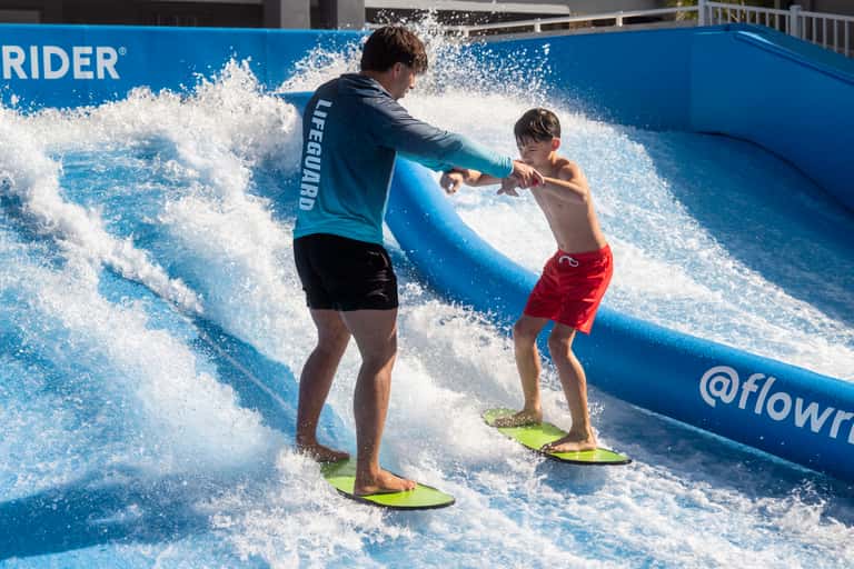 Instructor teaching young boy to surf on blue FlowRider wave with white water spray