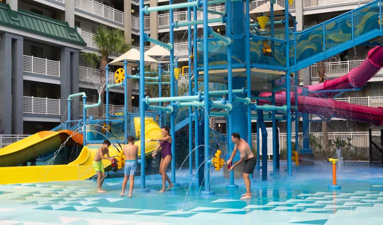 Group of children playing on blue water play structure with yellow and pink slides at resort splash pad