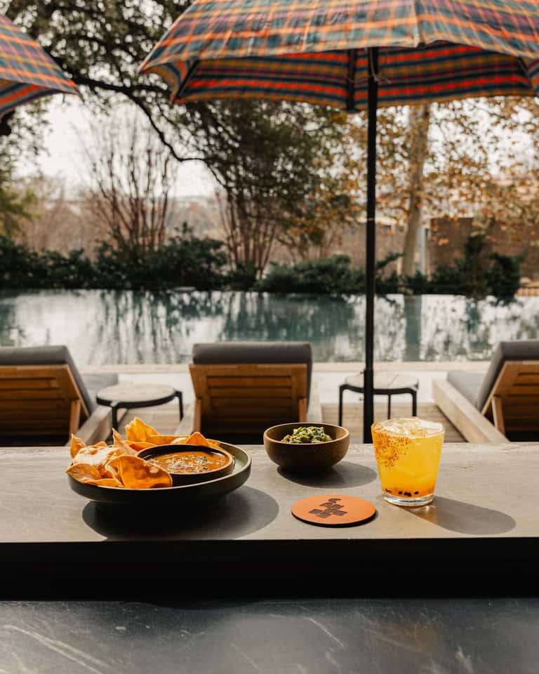 Outdoor dining table with food and beverage under checkered umbrella overlooking calm pool and landscape
