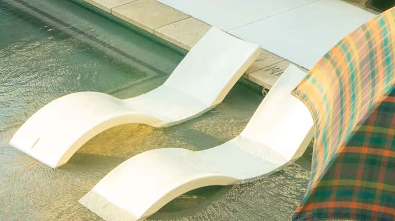 White curved lounge chairs partially submerged in shallow pool water with checkered umbrella nearby
