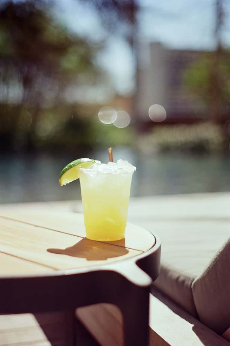 Refreshing yellow cocktail with lime wedge on wooden table overlooking blurred outdoor patio setting