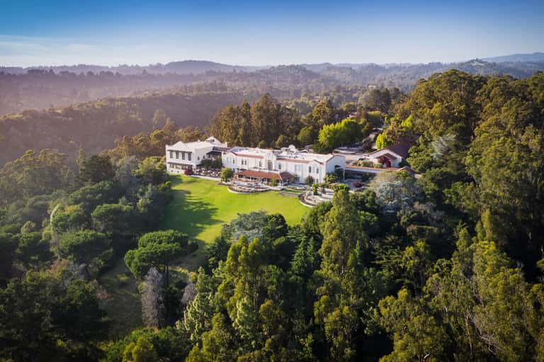 Aerial view of luxurious white mansion nestled in forested hillside with manicured lawns
