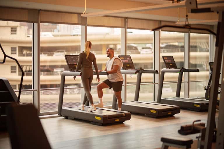 Modern gym with floor-to-ceiling windows overlooking city skyline, two people using treadmills