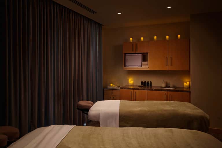 Luxury spa treatment room with two massage tables, wood cabinetry, and candlelit ambiance
