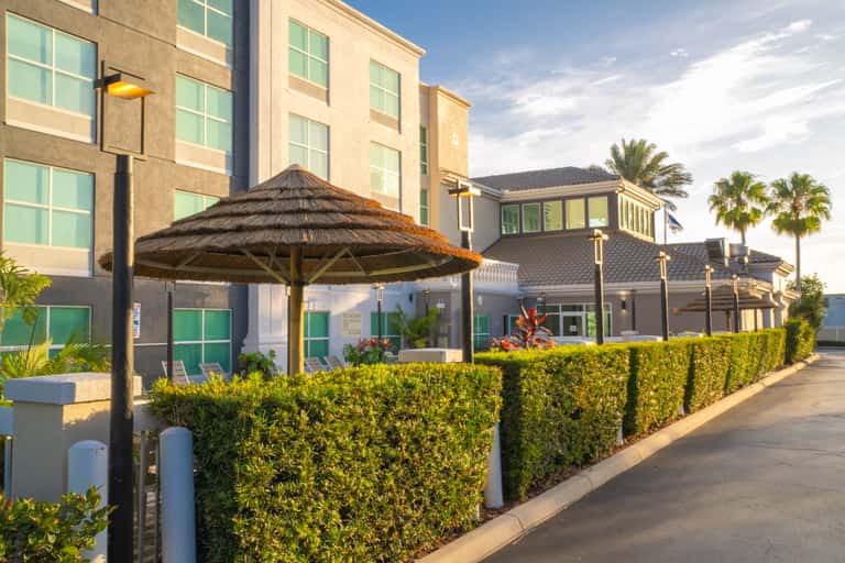 Modern hotel exterior with multi-story building, tropical landscaping, and thatched umbrella seating area