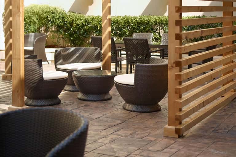 Patio seating area with woven chairs, white cushions, wooden pergola, and hedged privacy screening