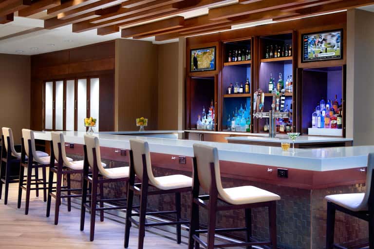Upscale bar lounge with white counter, wooden ceiling beams, illuminated liquor shelves, and cushioned bar stools