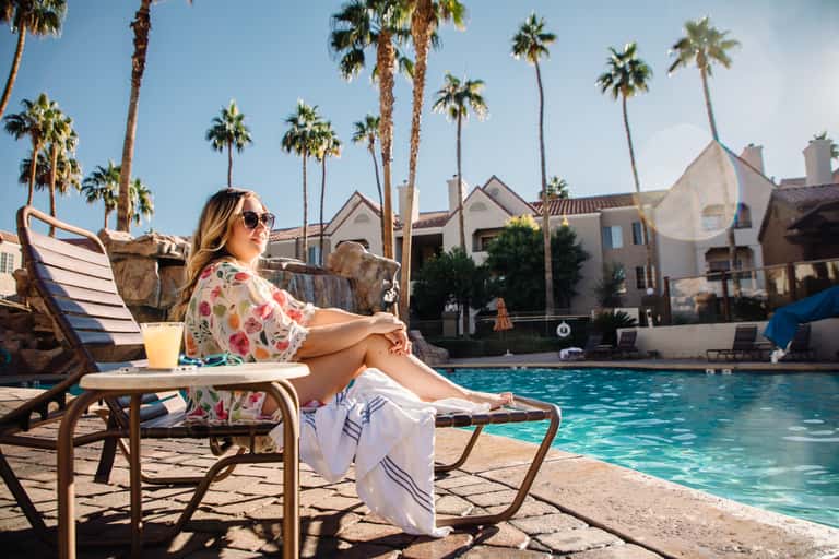 Woman in floral shirt relaxing poolside on lounge chair with palm trees and resort buildings in background