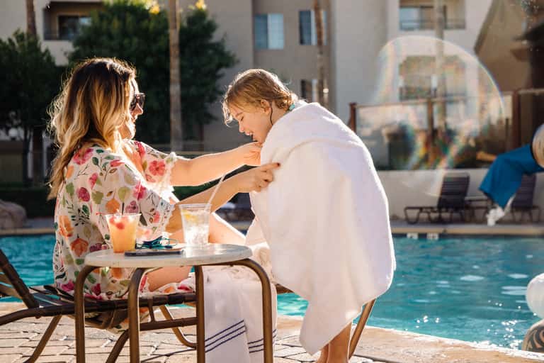 Woman wrapping child in white towel after swimming at resort pool during golden hour