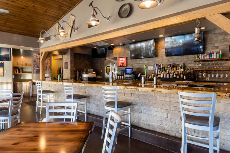 Modern bar interior with stone accent wall, wooden ceiling, white bar stools and flat-screen TVs