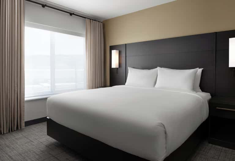 Modern hotel bedroom with king bed, black headboard, beige walls, and floor-to-ceiling windows with city views