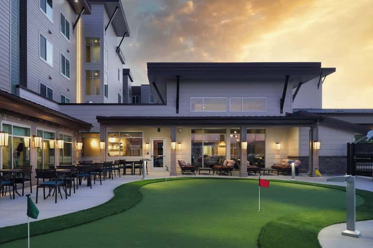 Upscale residential patio with dining area, lounge space, mini golf course, and illuminated building at dusk