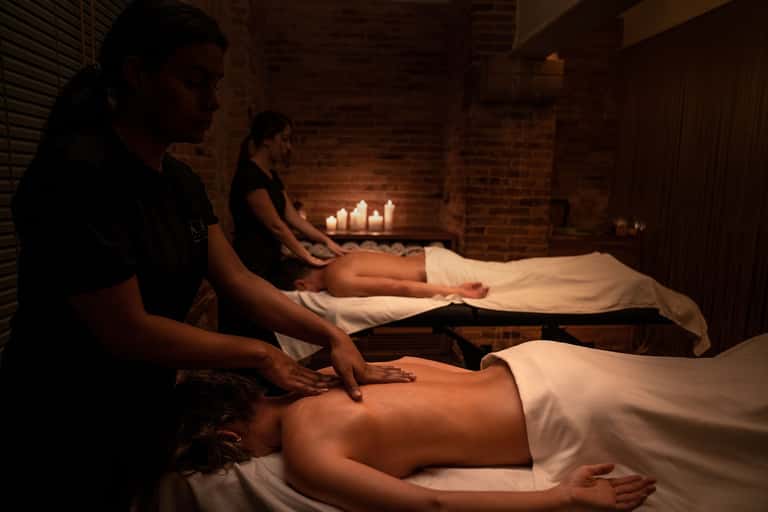 Two massage therapists performing treatments on clients in candlelit spa room with brick walls