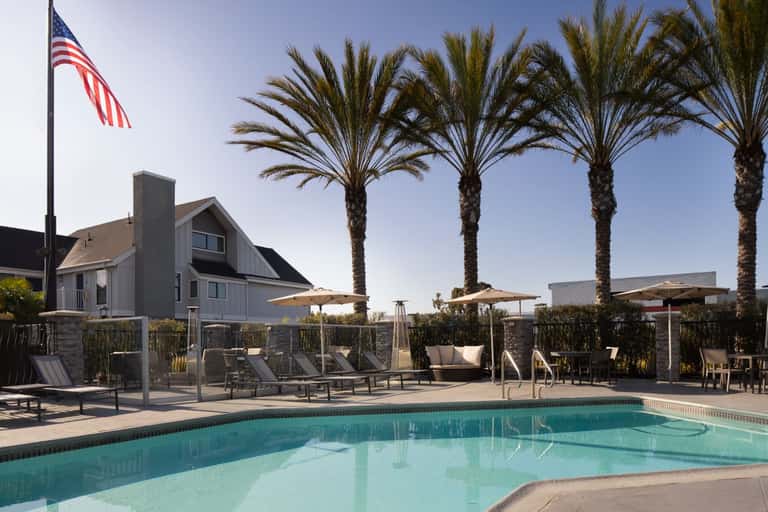 Coastal pool area with palm trees, umbrella shade, lounge chairs, and modern cottage-style building