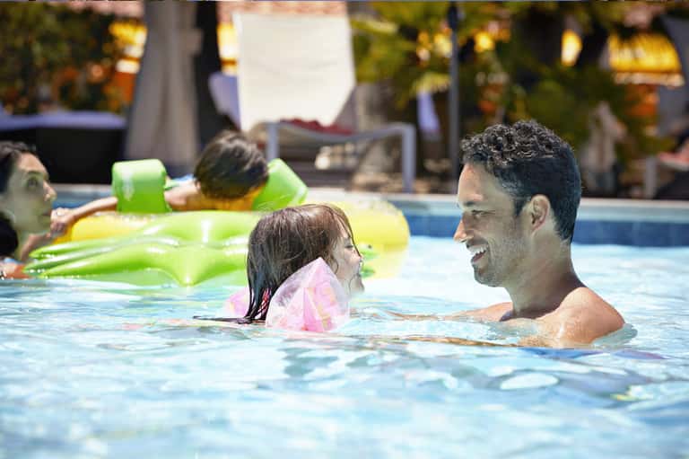 Father and young daughter swimming together in outdoor resort pool with floating toys during daytime
