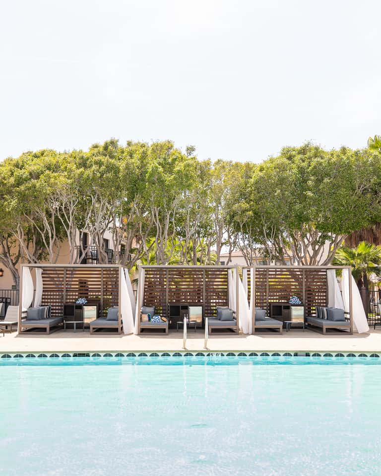 Resort poolside seating area with individual cabanas featuring wooden screens, cushioned lounges, and flowering plants along the pool deck