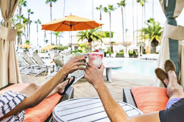 Guests toasting with colorful cocktails by the pool with orange umbrellas and palm trees in background