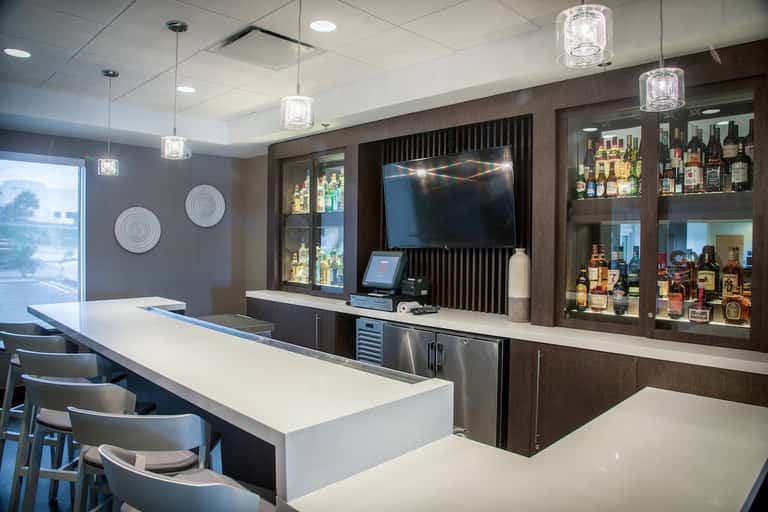 Modern hotel bar with dark wood cabinetry, lit liquor display, and white countertop