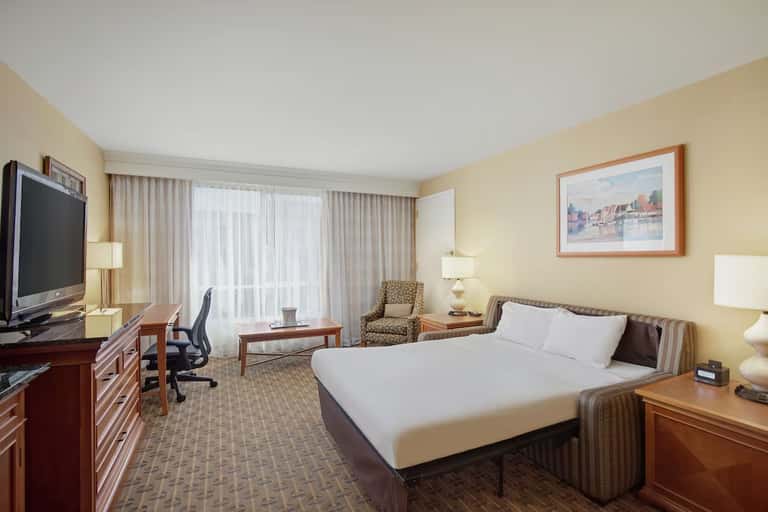 Hotel room with single bed, work desk, flat-screen TV, and seating area overlooking balcony