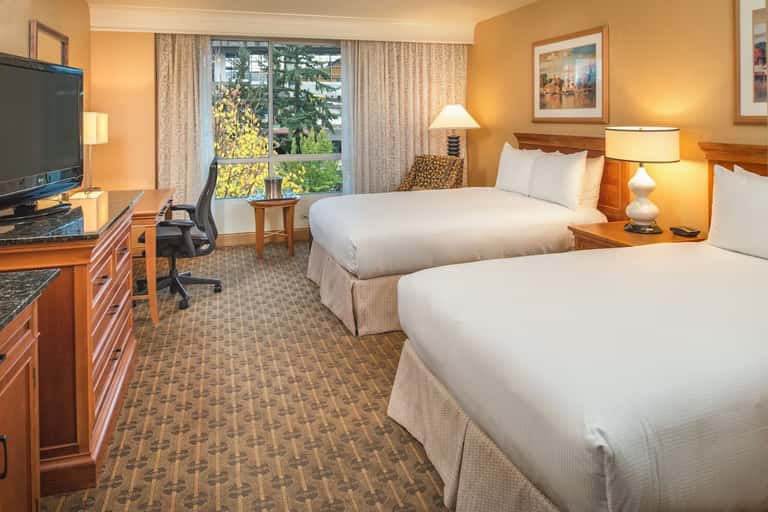 Hotel guest room with two queen beds, wooden furnishings, work desk, and large windows overlooking trees