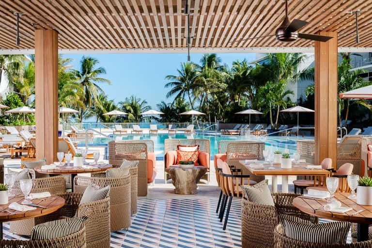 Luxury poolside dining area with striped wooden pergola, woven chairs, and ocean-view resort in background