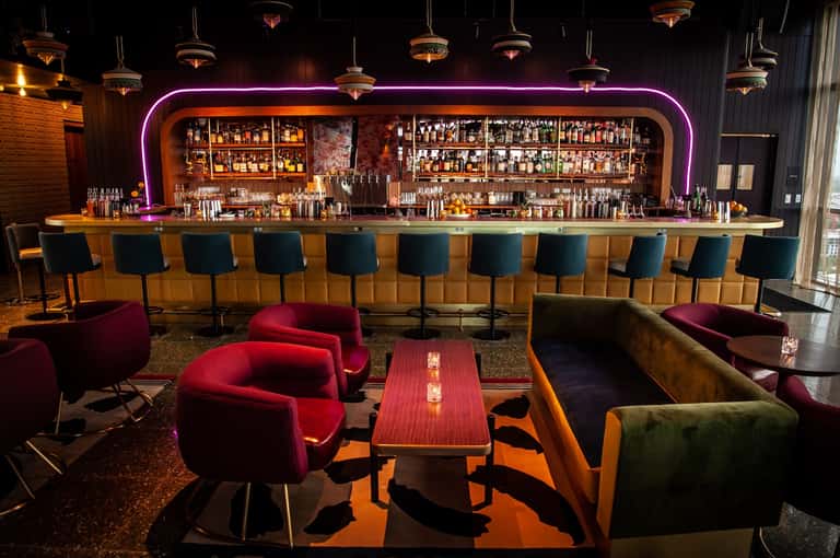 Upscale bar lounge with neon pink signage, burgundy seating, wood paneling, and extensive liquor shelves