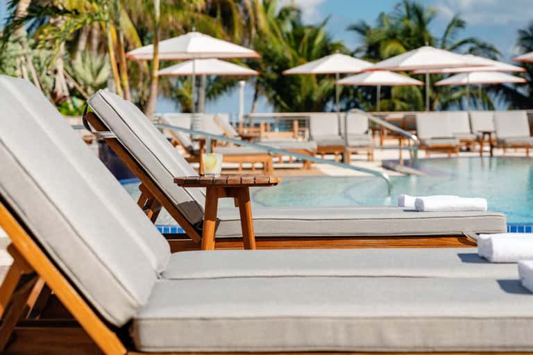 Lounge chair with cocktail by resort pool surrounded by white umbrellas and palm trees