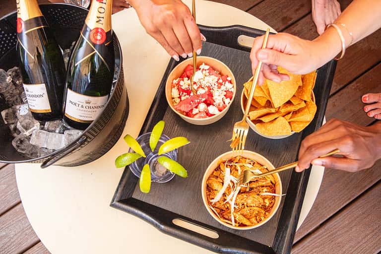 Overhead view of champagne bottles, gourmet appetizers, and lime garnishes on wooden poolside table
