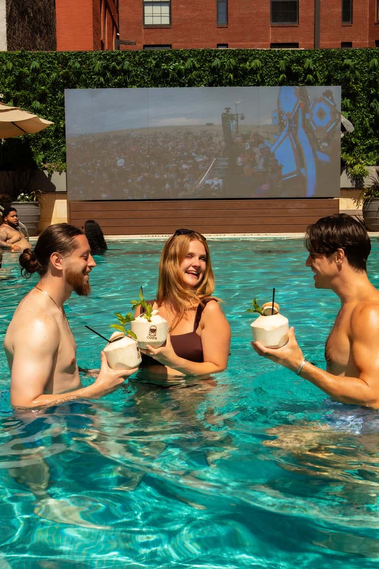 Guests in turquoise pool holding coconut drinks with outdoor movie screen and brick building backdrop