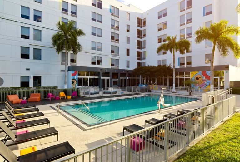 Resort pool area with colorful lounge chairs, palm trees, and modern white apartment building