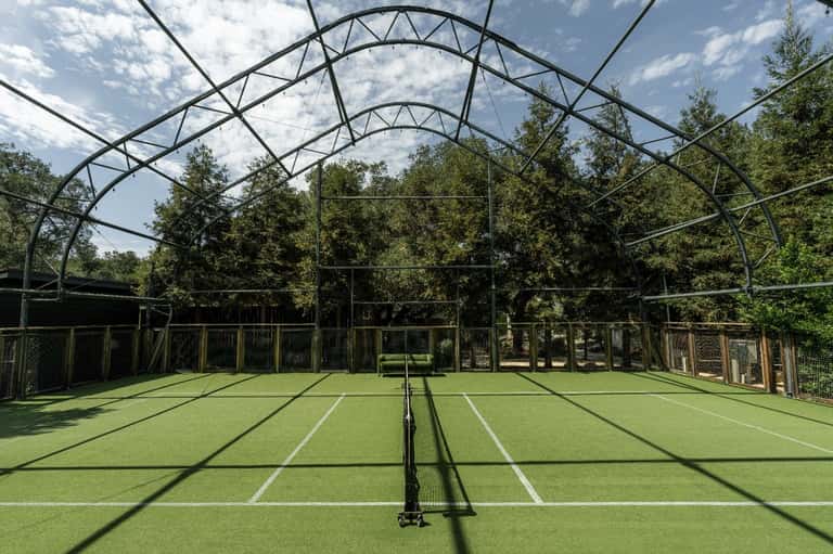 Indoor tennis court with white markings, metal framework roof, and natural lighting