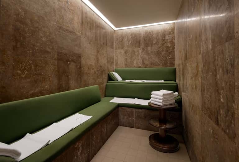 Spa relaxation room with green cushioned benches, travertine walls, and ambient ceiling lighting