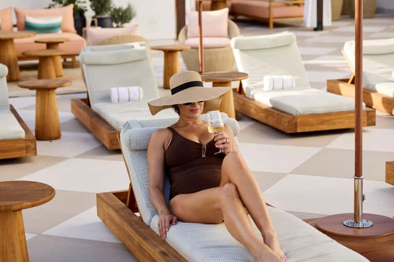 Woman in wide-brimmed hat relaxing on wooden lounge chair with white cushion, holding champagne glass at resort pool deck