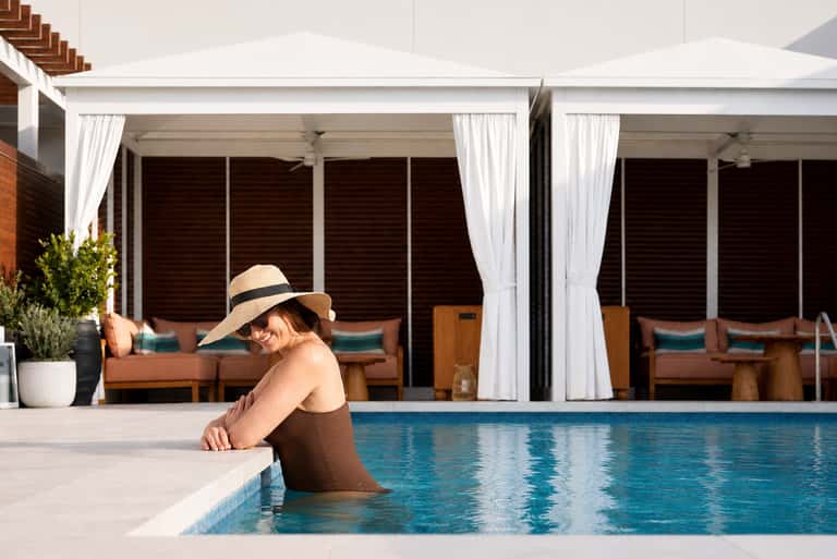 Woman in hat swimming in indoor pool with white cabanas and lounge chairs with brown cushions visible in background