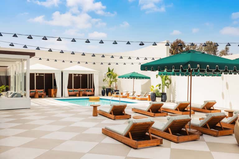 Modern resort pool area with white pergolas, green umbrellas, wooden lounge chairs with cushions, and contemporary architecture